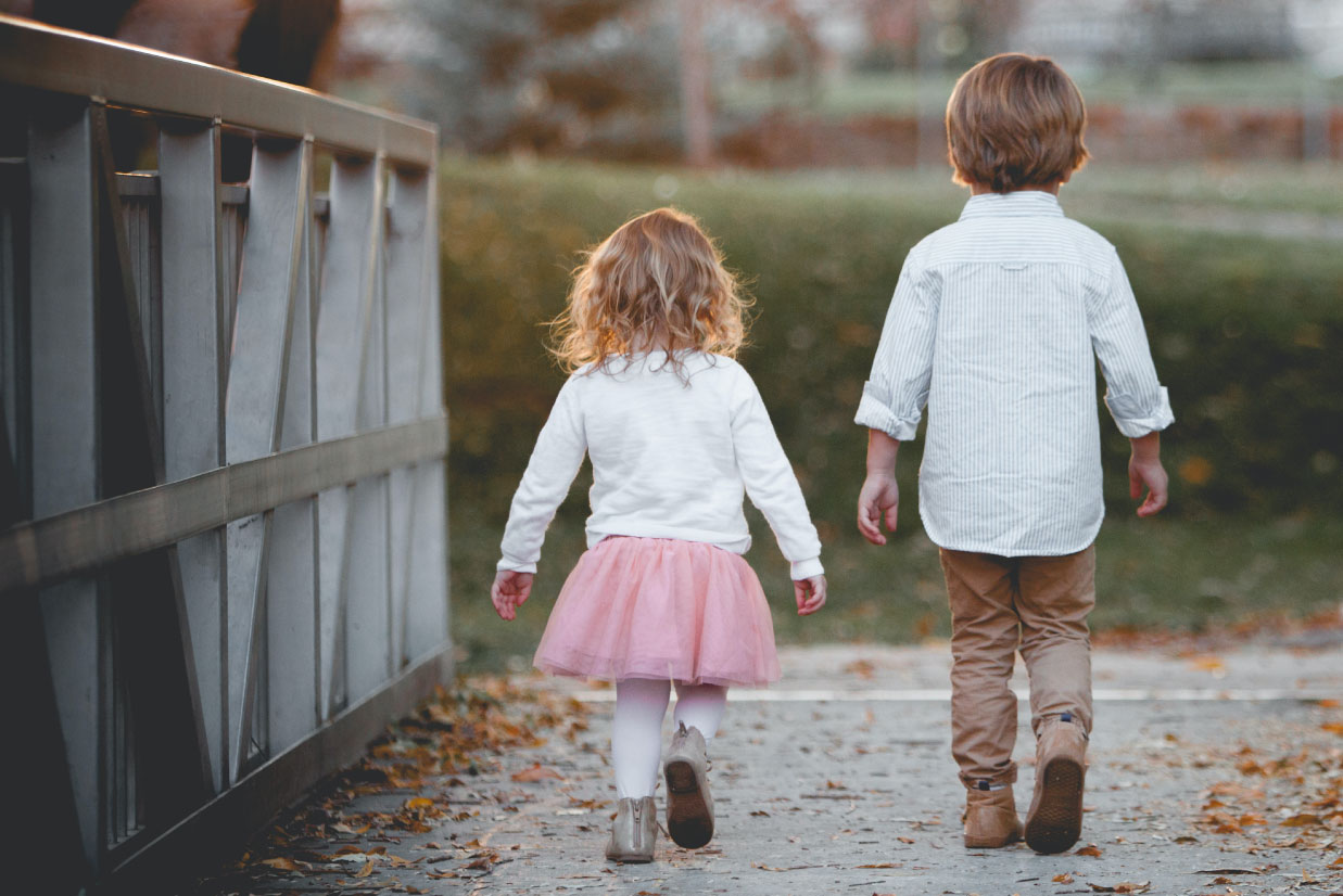 Two children walking on a path, a girl in a pink tutu and a boy in a striped shirt, surrounded by autumn leaves and a bridge in the background.