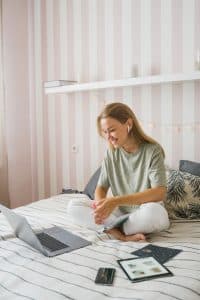 Woman sitting on a bed with a laptop, tablet, and smartphone, engaged in online activities, reflecting a comfortable home environment suitable for remote work or home buying consultations.