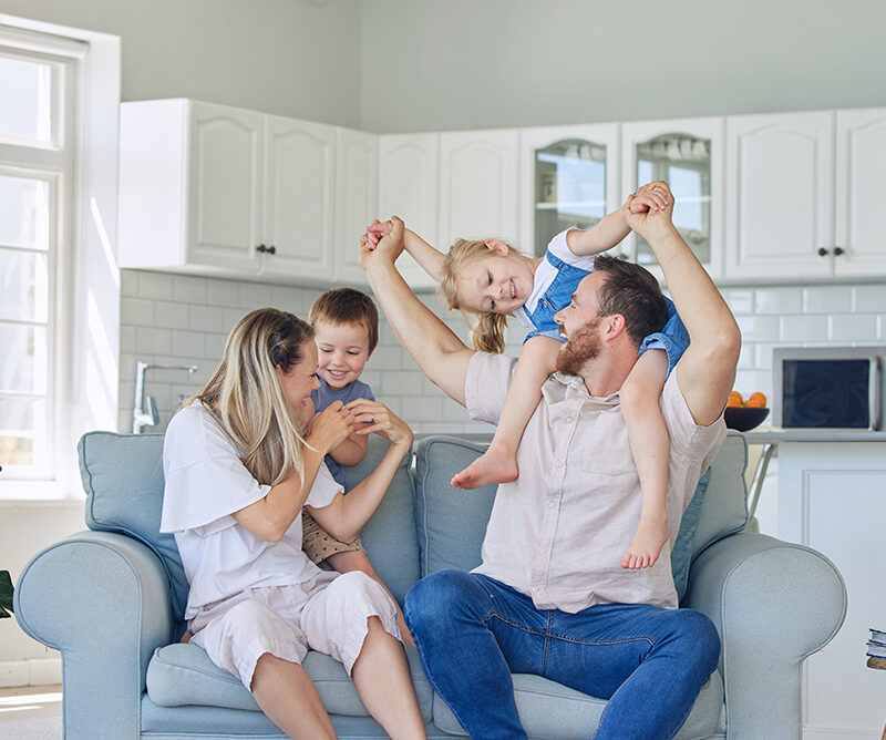 Family of four sitting on a sofa, smiling and enjoying time together in a bright, modern living room, illustrating the joy of homeownership and family life relevant to USDA home loan benefits.