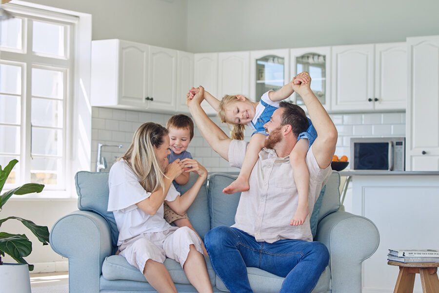 Family of four sitting on a sofa, smiling and enjoying quality time together, in a cozy living room setting, reflecting themes of home and family in mortgage discussions.