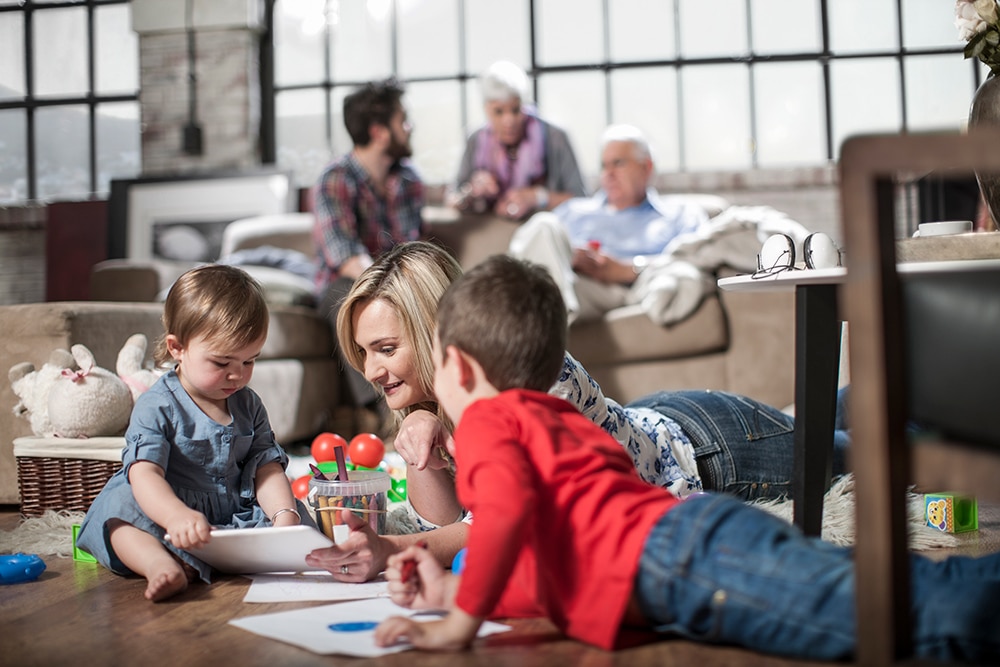 Mother engaging with children drawing at home, family atmosphere, toys scattered, bright interior space, promoting family values and comfort.