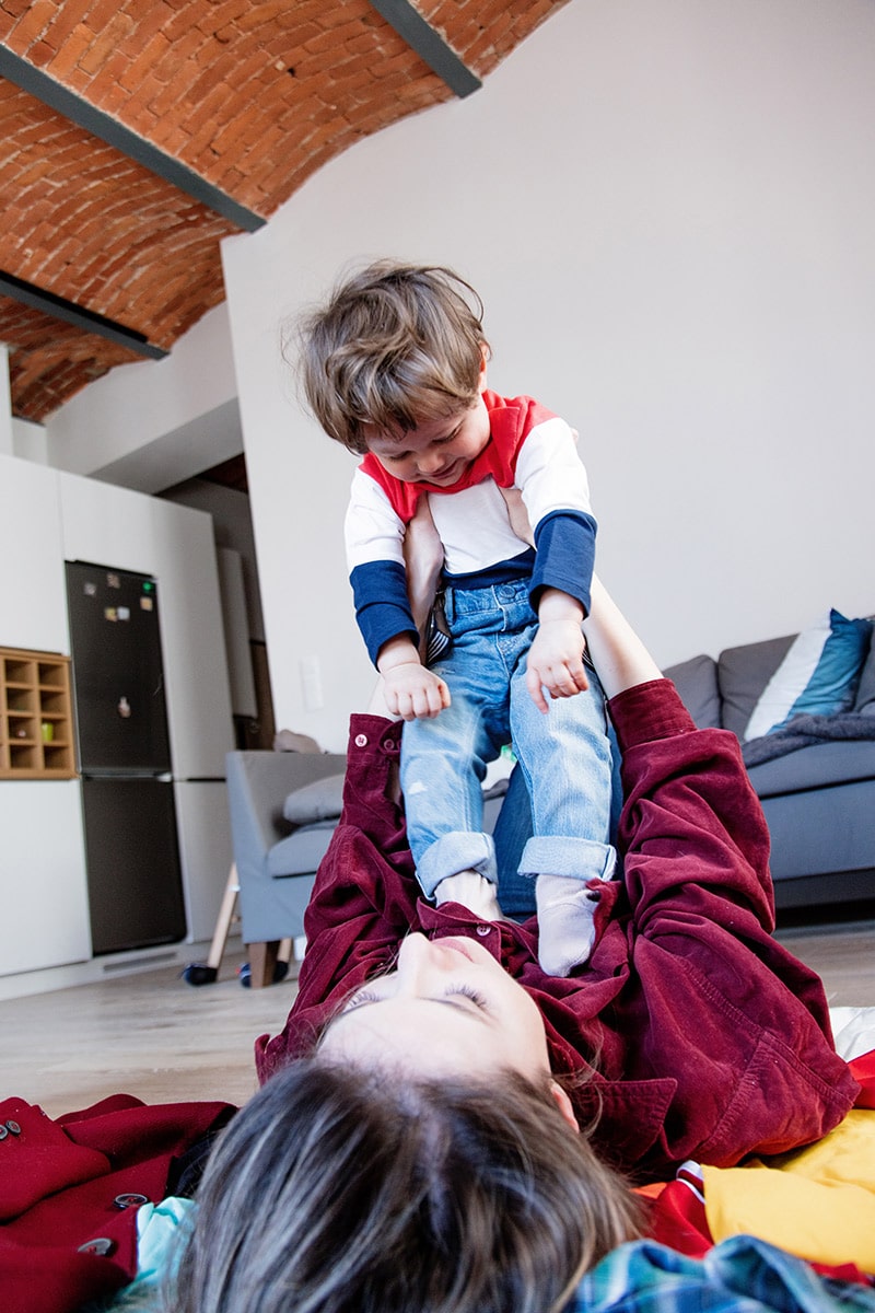 Mother and son playing together on the floor, child joyfully lifted above, in a cozy living room setting, highlighting family bonding and playful interaction.