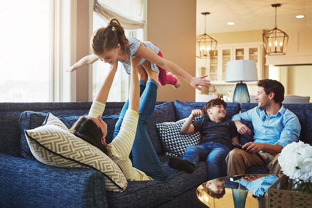 Family enjoying playful moments at home, mother lifting daughter while two boys laugh on the couch, emphasizing joy and togetherness in a cozy living space.