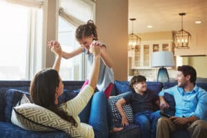 Family enjoying quality time together in a cozy living room, with a mother playfully lifting a daughter while a son and father watch, highlighting the joys of family life and emotional connection.